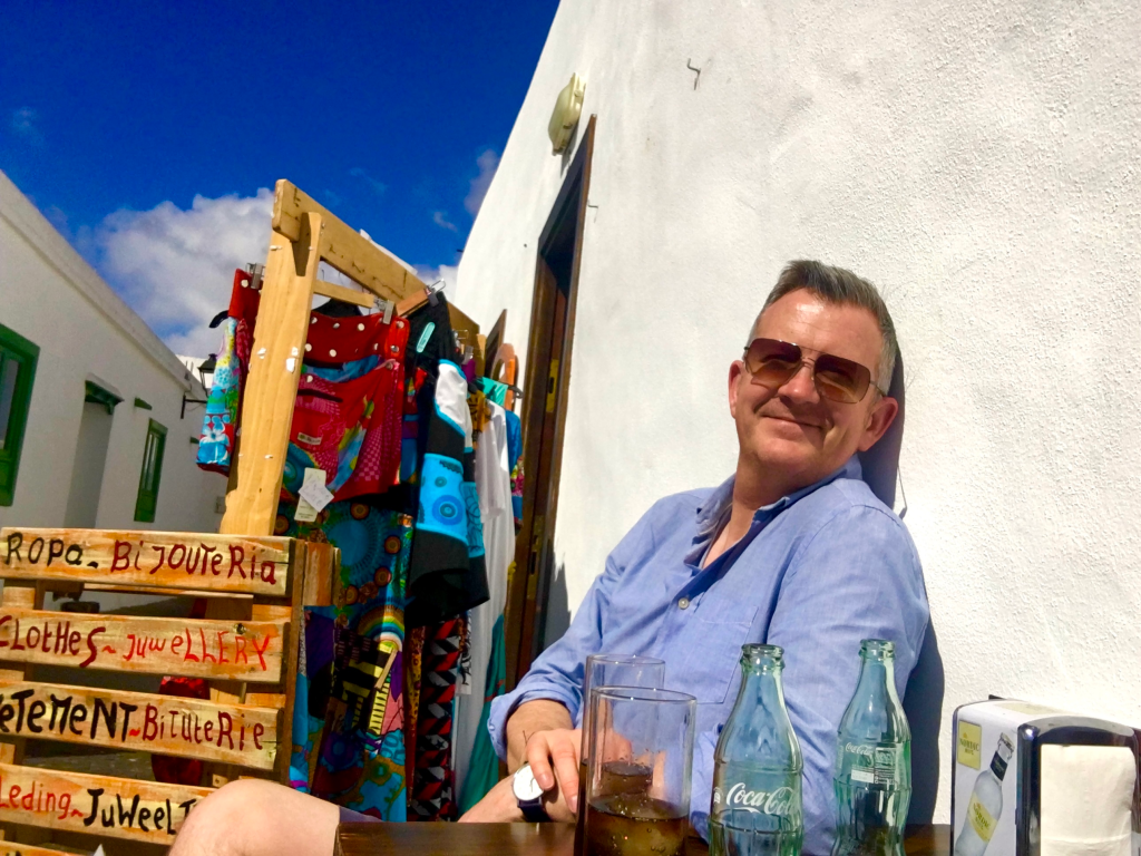 Author sitting in a sunny side street, white wall behind him, colourful basic signs to the left.
